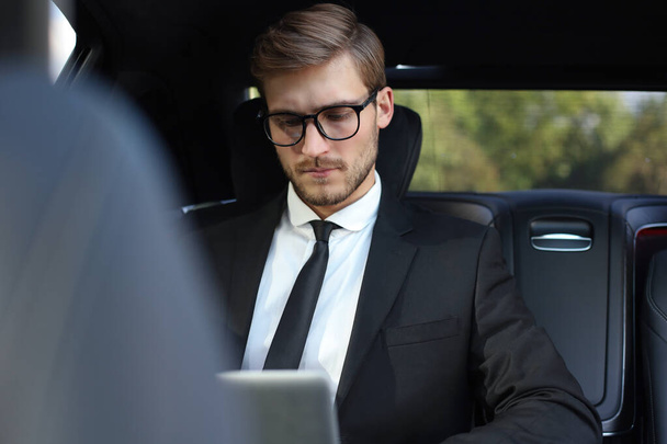 Guapo empresario confiado en traje hablando en el teléfono inteligente y trabajando con el ordenador portátil mientras está sentado en el coche
 - Foto, imagen