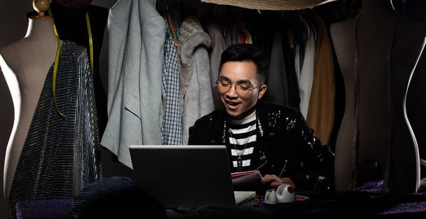 Black Sequin jas van Aziatische Fashion Designer Man glimlach blij om te krijgen order verkoop groei op Notebook computer. gay kleermaker ontwerpt nieuwe trend van collectie naar doek rek achtergronden, lage sleutel kopieerruimte - Foto, afbeelding