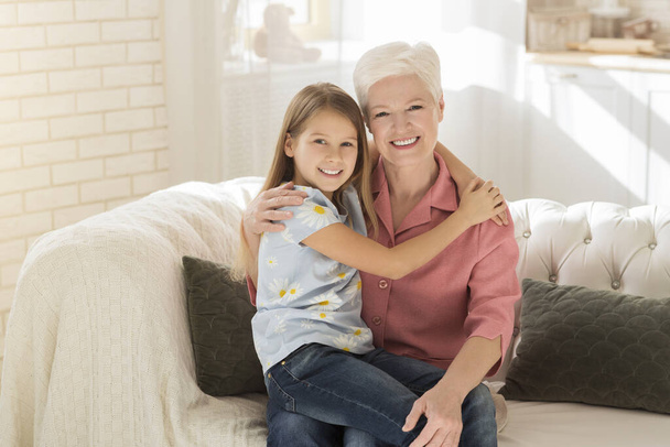 Friendship between generations. Cute kid with her granny hugging in sunlit room - Photo, Image