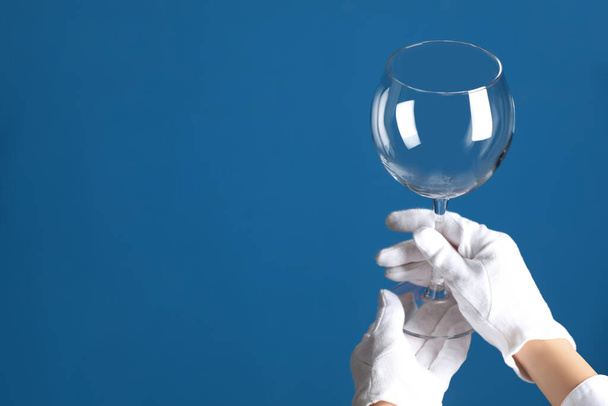 Person in white gloves checking cleanliness of glass on blue background, closeup. Space for text - Photo, image