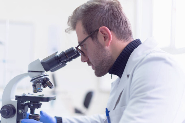 Νεαρός αρσενικός επιστήμονας που κοιτάζει μέσα από ένα μικροσκόπιο σε ένα εργαστήριο κάνοντας έρευνα, μικροβιολογική ανάλυση, ιατρική. - Φωτογραφία, εικόνα