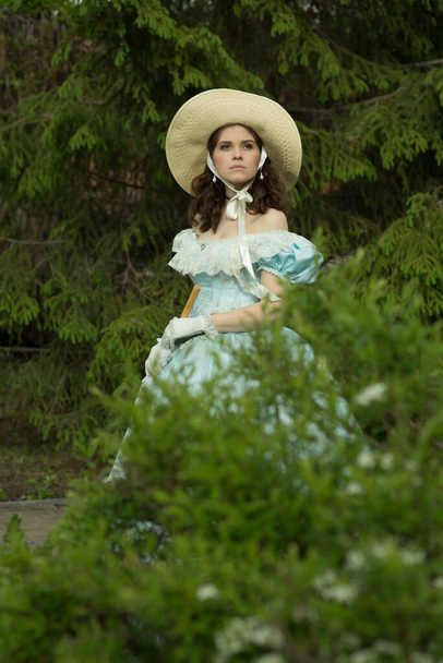 Ένα όμορφο κορίτσι σε ένα πρησμένο μπλε φόρεμα και καπέλο, κρατώντας μια ομπρέλα δαντέλας σε διπλωμένη κατάσταση, κοιτάζει αλλού. Σε πρώτο πλάνο είναι ένα κλαδί από ένα δέντρο. Ιστορική ανακατασκευή - Φωτογραφία, εικόνα