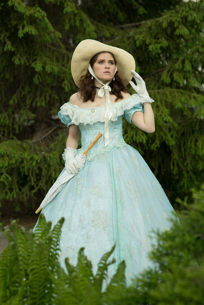A beautiful girl in a puffy blue dress and hat, holding a lace umbrella in a folded state, looks up. Historical reconstruction - Photo, Image