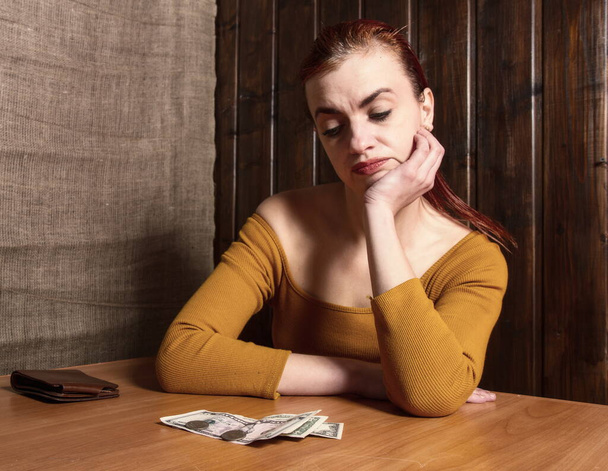 triest meisje in een nogal arm interieur kijkt naar de balans van geld in de voorkant van haar - Foto, afbeelding
