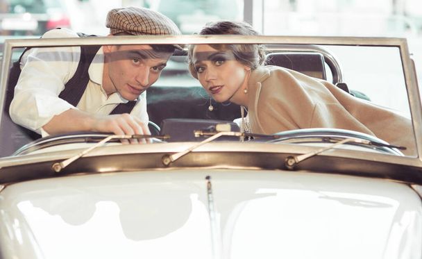 Una joven pareja está sentada en un coche retro. Un hombre enseña a una mujer a conducir, y le dice algo. Reconstrucción histórica
 - Foto, Imagen