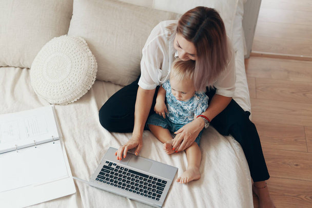 Jonge moeder met haar kind die thuis op laptop werkt in de slaapkamer. Multi-tasking, freelance en moederschap concept - Foto, afbeelding