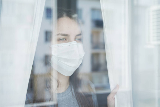 Young Woman in Face Mask Looking out the Window. Staying Home in Quarantine. - Zdjęcie, obraz