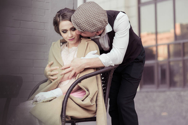 Um jovem abraça uma mulher que está sentada em uma cadeira de vime. Roupas em estilo retro. Reconstrução histórica
 - Foto, Imagem