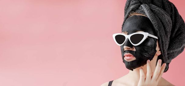 Young woman in glasses appling black cosmetic fabric facial mask on pink background. Face peeling mask with charcoal, spa beauty treatment, skincare, cosmetology. Close up. - Foto, afbeelding
