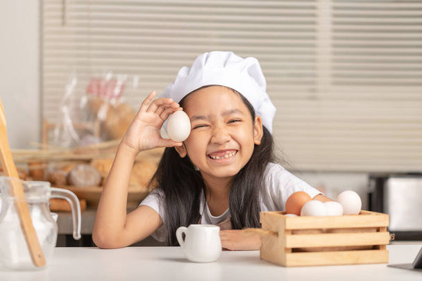 Asian little girl wearing a white chef hat is holding a duck egg and smiling. Eggs in a wooden basket in the kitchen. - Photo, Image