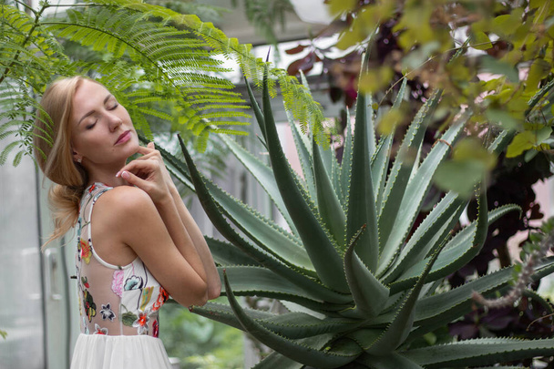 Blond europees jong vrouw tussen groene palmen en cactussen in tropische tuin in witte jurk en lang haar - Foto, afbeelding