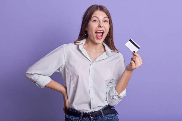 Indoor studio portrait of magnetic attractive young female posing isolated over lilac background, holding credit card, opening mouth, keeping one hand on waist, making credit payments successfully. - Foto, Imagem