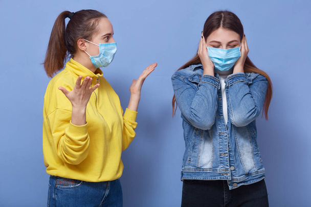 Image of two young women standing isolated over blue background, lady wearing denim jacket covers ears, female dresses yellow shirt making helpless gesture, girls in medical mask, to protect covid 19. - Foto, immagini
