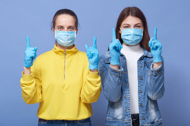 Covid 19, coronavirus concept. Young Caucasian women wearing medical mask and gloves, casual outfits, standing and pointing up with index fingers, beautiful ladies trying to avoid virus infection. - 写真・画像
