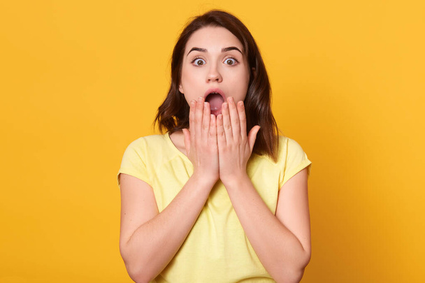 Close up portrait of astonished woman with widely open mouth, being amazement, stands with big eyes, looking at camera, stunned shocked face isolated over yellow background. People emotions concept. - Photo, image