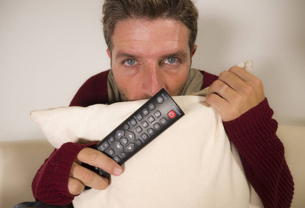 home lifestyle portrait of young attractive scared and stressed man at living room couch holding TV remote watching horror movie or scary news on television about covid-19 virus outbreak  - Foto, Bild