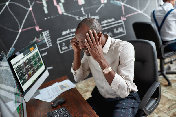 Feeling bad. Portrait of stylish african businessman, trader sitting by desk in front of multiple monitors. He is getting angry while looking at current graphs - Fotoğraf, Görsel