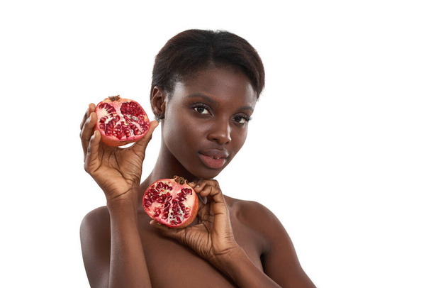 Add pomegranate to your skincare. Portrait of beautiful young african woman holding two pieces of fresh juicy pomegranate fruit and looking at camera while standing against white background - Fotografie, Obrázek