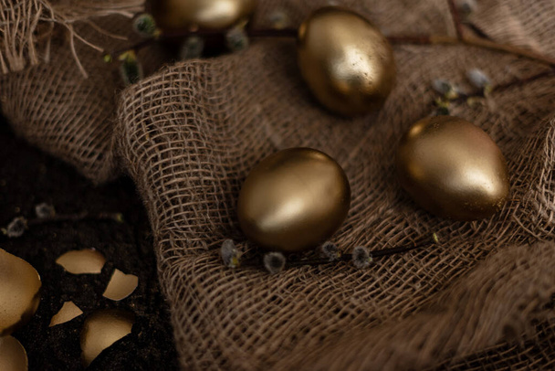 Golden easter eggs are scattered on a dark background with dry green branches. eggs in a golden bowl. - Фото, зображення
