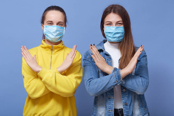 明るい服と医療使い捨てマスクを身に着けている2人の若い女性の屋内ショットは、青の背景に手でジェスチャーを停止表示します。インフルエンザ、コロナウイルスまたは感染症の概念. - 写真・画像