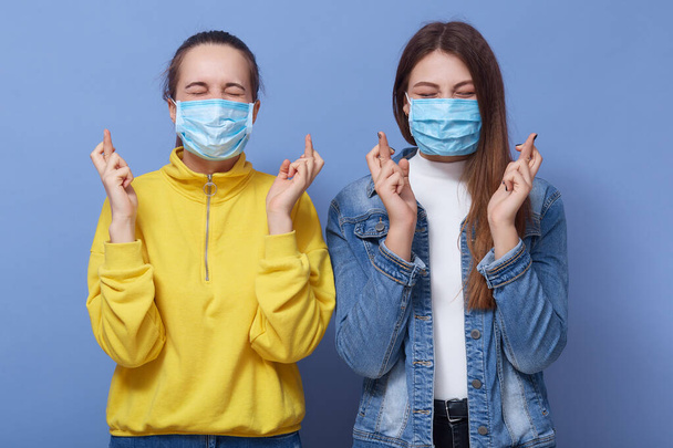 Portrait of two young women with cross fingers, wearing protective face mask, preventing virus infection, ladies dress yellow sweatshirt anf denim jacket, posing isolated over blue background. - 写真・画像
