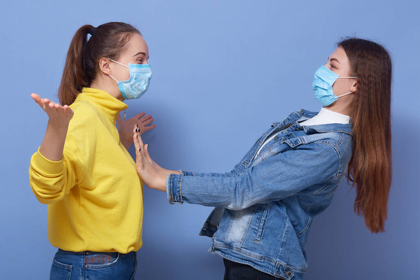 Adorable female don't want to contact with her friend wearing yellow shirt and mask, trying to avoid pandemic disease, Corona virus, covid-19. Lady looks scared at young girl with spreading hands. - Фото, изображение