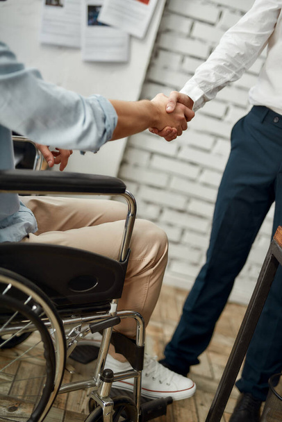 Vystřižený pohled na pracovníka mužské kanceláře na invalidním vozíku, jak si potřásá rukou se svým kolegou, když pracuje společně v kreativní kanceláři. Koncept zdravotního postižení - Fotografie, Obrázek