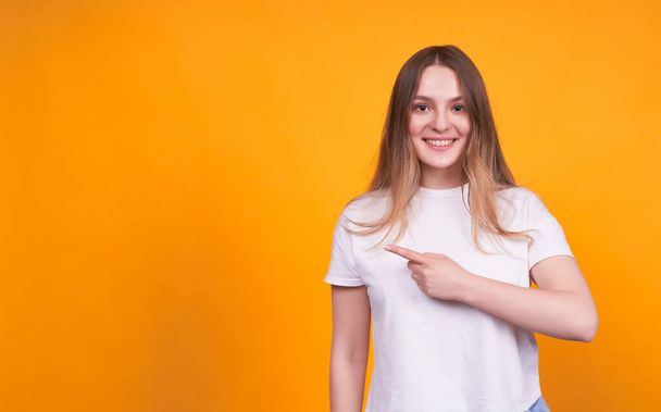 Young cute and pretty girl in a white T-shirt on a yellow isolated background, smiling, pointing her finger to the left to space for your advertisement or text. - 写真・画像
