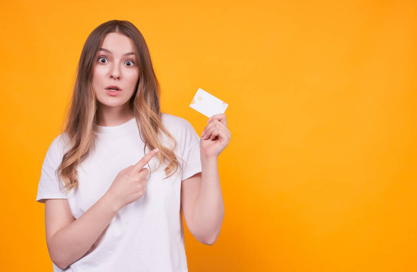 Portrait of a happy cute young girl with a funny face showing a index finger on a plastic credit card, not understanding where spent money, isolated on a yellow background.Copy space. - Фото, зображення
