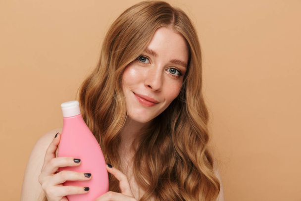 Portrait closeup of gorgeous young half-naked woman holding hair treatment bottle isolated over beige background - Foto, Imagem