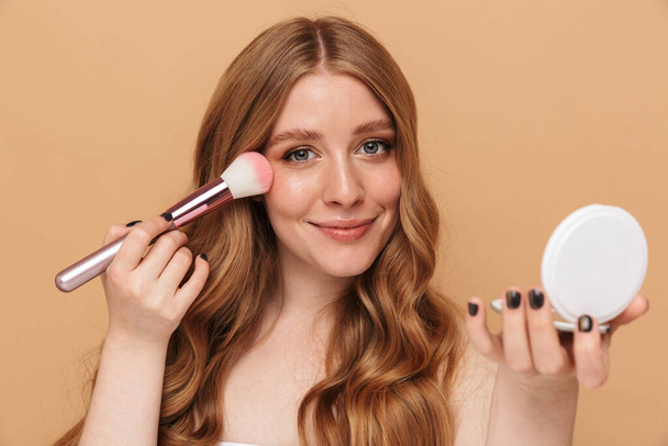 Portrait closeup of gorgeous young half-naked woman with long hair applying makeup with face brush isolated over beige background - Foto, afbeelding