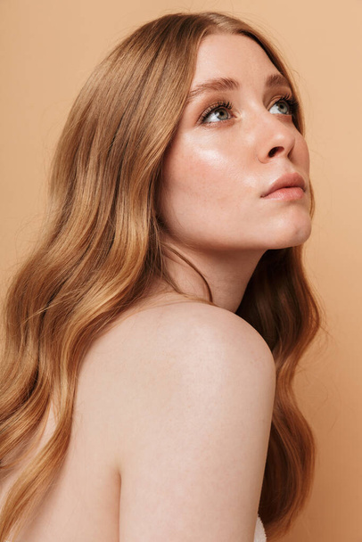 Portrait closeup of gorgeous young half-naked woman with long auburn hair looking aside at copyspace isolated over beige background - Foto, Imagem