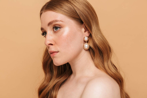 Portrait closeup of gorgeous young half-naked woman with long auburn hair looking aside at copyspace isolated over beige background - Foto, afbeelding