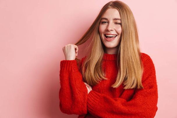 Photo of happy beautiful woman in red sweater smiling and winking while posing isolated over pink wall - Photo, image