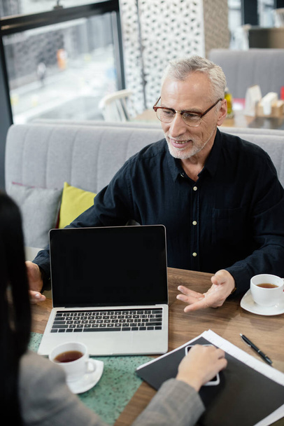 Улыбающийся бизнесмен разговаривает с деловой женщиной и сидит рядом с ноутбуком во время деловой встречи
  - Фото, изображение