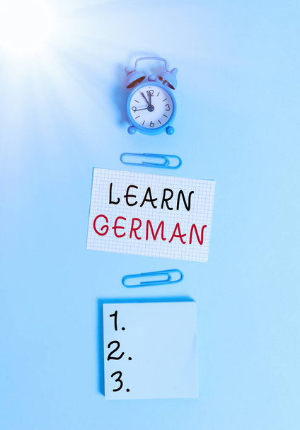 Text sign showing Learn German. Conceptual photo get knowledge or skill in speaking and writing German language Alarm clock squared blank paper sheet notepad clips colored background. - Photo, Image