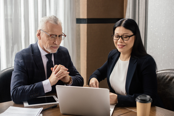 businessman and smiling asian businesswoman looking at laptop during business meeting  - Photo, Image
