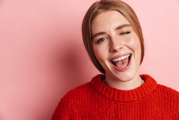 Photo of happy beautiful woman in red sweater smiling and winking while posing isolated over pink wall - Valokuva, kuva