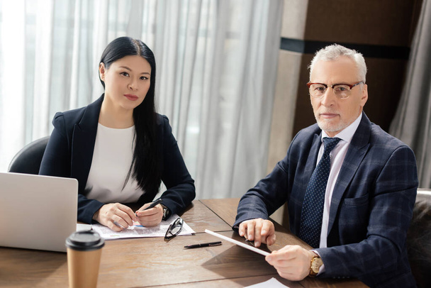 businessman and asian businesswoman looking at camera and sitting at table - Fotografie, Obrázek