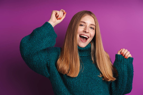 Photo of cheerful caucasian woman in green sweater smiling and dancing isolated over violet wall - 写真・画像