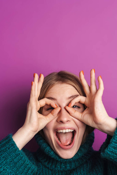 Photo of excited caucasian woman in green sweater making fun with fingers isolated over violet wall - Zdjęcie, obraz
