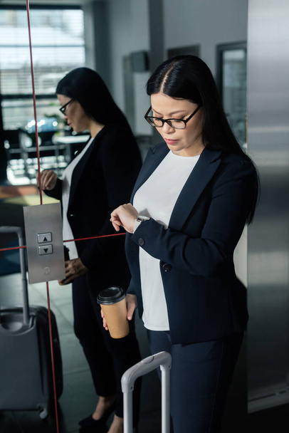 азиатская деловая женщина с бумажной чашкой, глядя на наручные часы и стоя рядом с лифтом
   - Фото, изображение