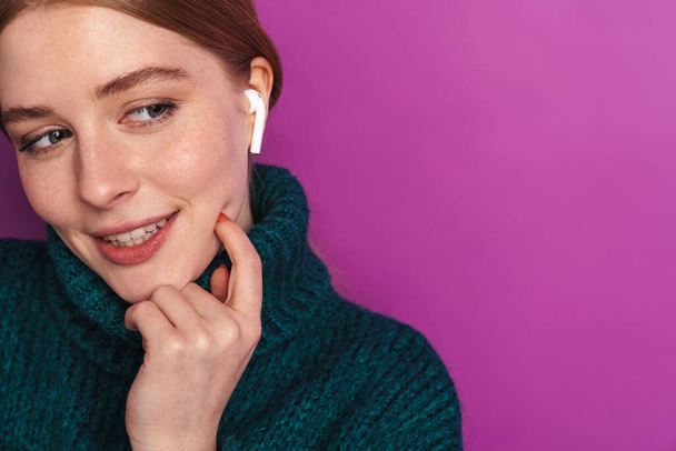 Photo of smiling caucasian woman in green sweater using wireless earphone isolated over violet wall - Foto, imagen