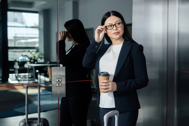 азиатская деловая женщина с бумажной чашкой глядя в сторону и стоя рядом с лифтом
   - Фото, изображение
