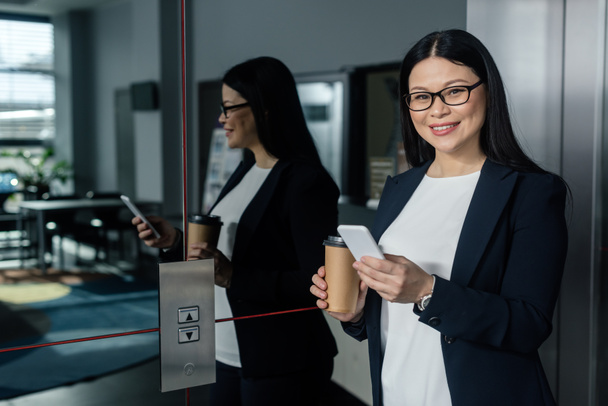 Lächelnde asiatische Geschäftsfrau mit Pappbecher, Smartphone und Blick in die Kamera  - Foto, Bild