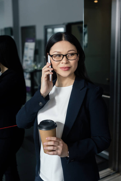 Lächelnde asiatische Geschäftsfrau hält Pappbecher in der Hand und spricht auf dem Smartphone  - Foto, Bild
