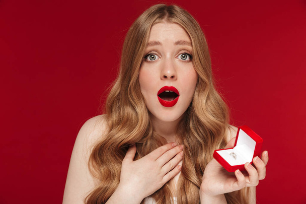 Image of young shocked woman posing isolated over red wall background with bright red lipstick holding present box with ring. - Photo, Image