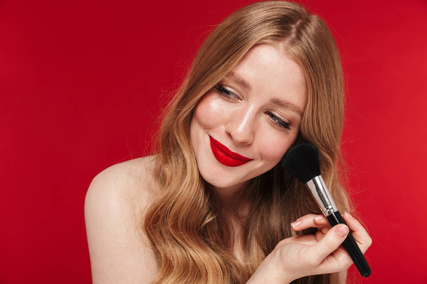 Image of a beautiful smiling pretty caucasian woman posing isolated over red wall background with bright red lipstick apply makeup with brush. - Foto, imagen