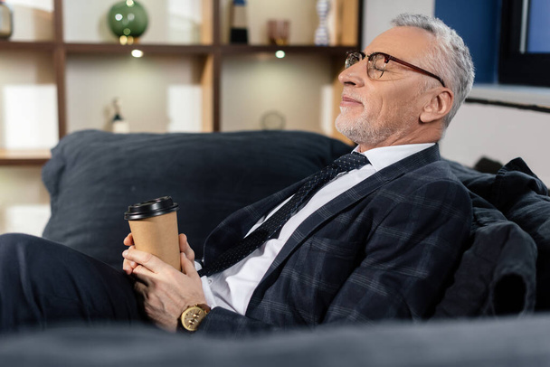 side view of smiling businessman with closed eyes sitting on sofa and holding paper cup  - Foto, Imagem