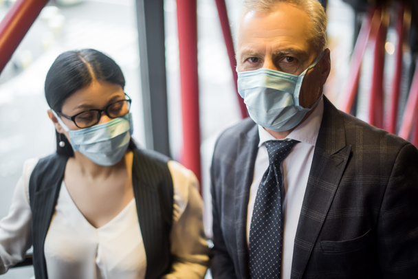 selective focus of businessman and asian businesswoman in medical masks looking at camera  - Photo, Image
